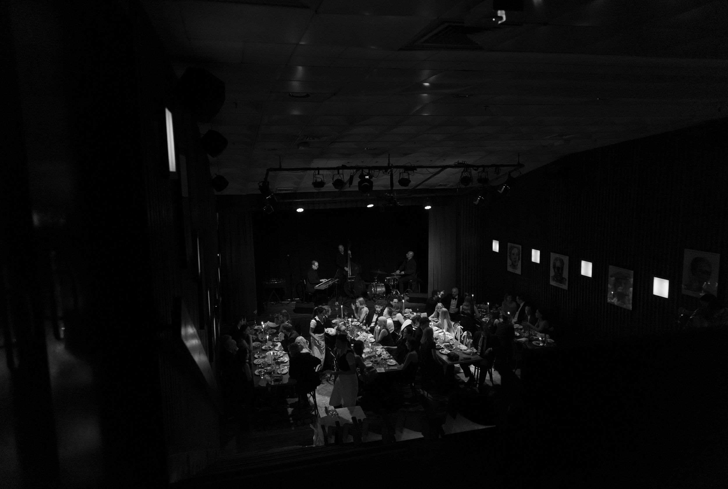 Black and white aerial view of a dimly lit banquet hall filled with guests seated at long dining tables.