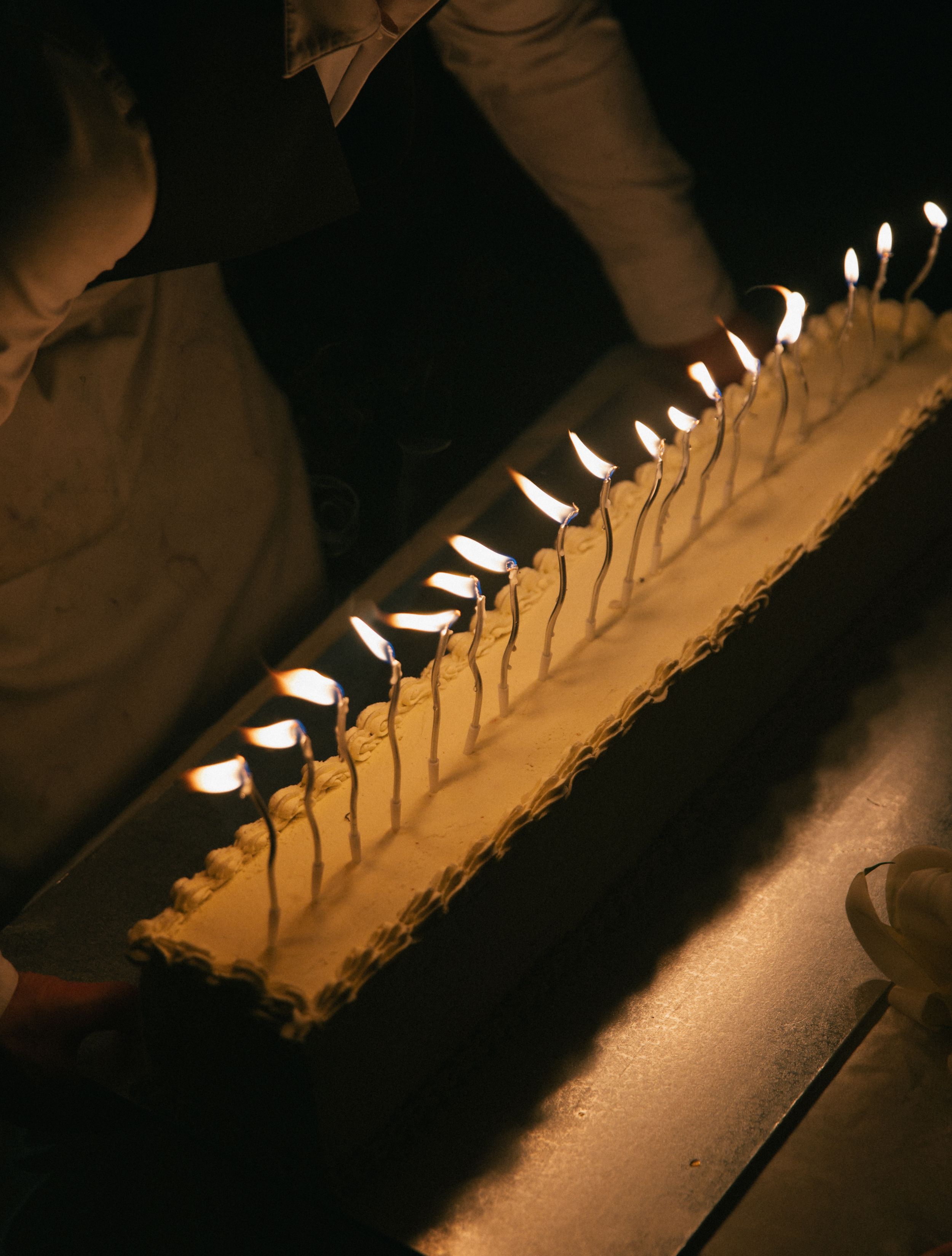 Long rectangular birthday cake with many lit candles glowing warmly in a dark room, being lit by two people.