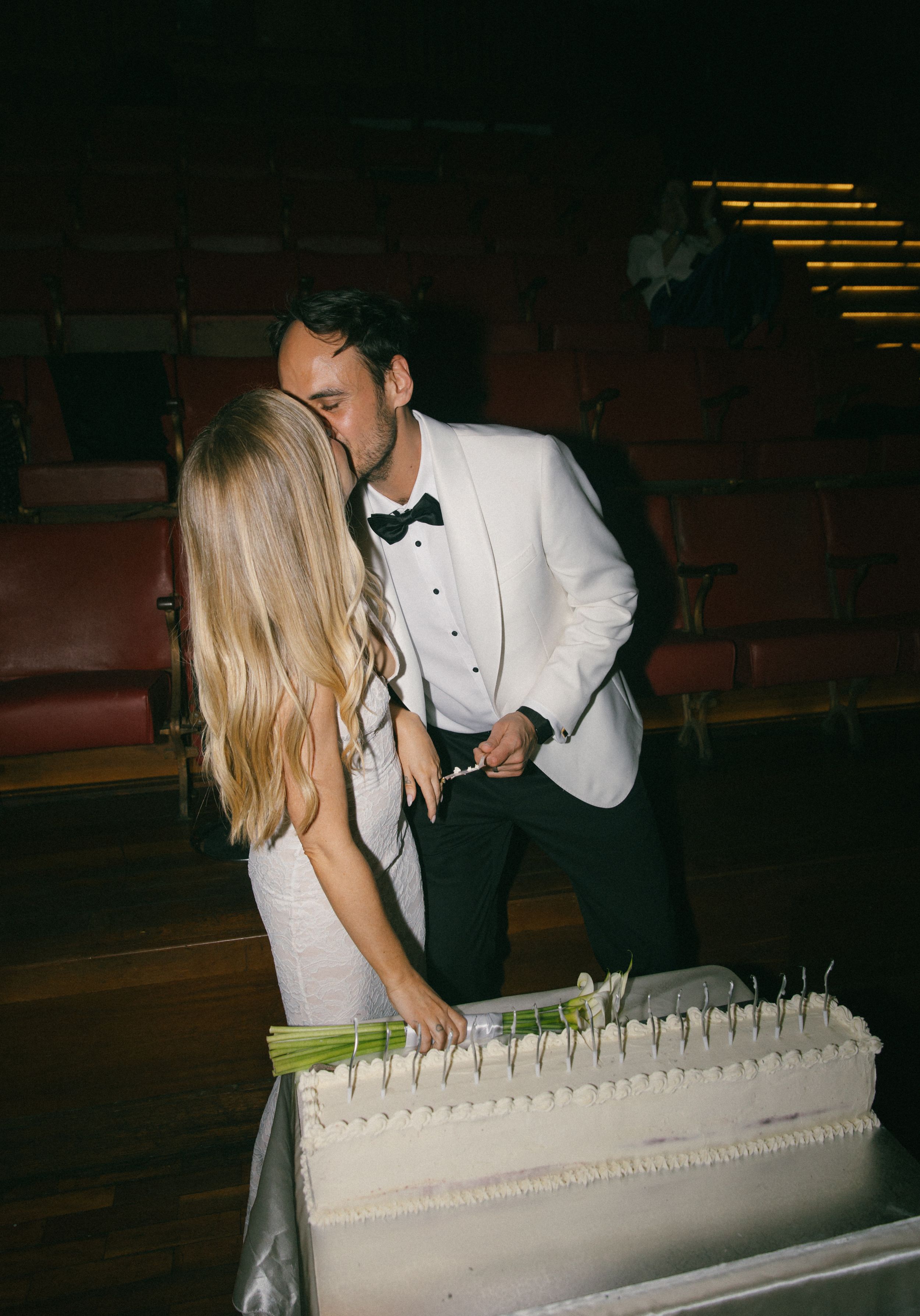 Couple sharing a kiss while cutting their white wedding cake, groom in white tuxedo jacket, bride in white gown.
