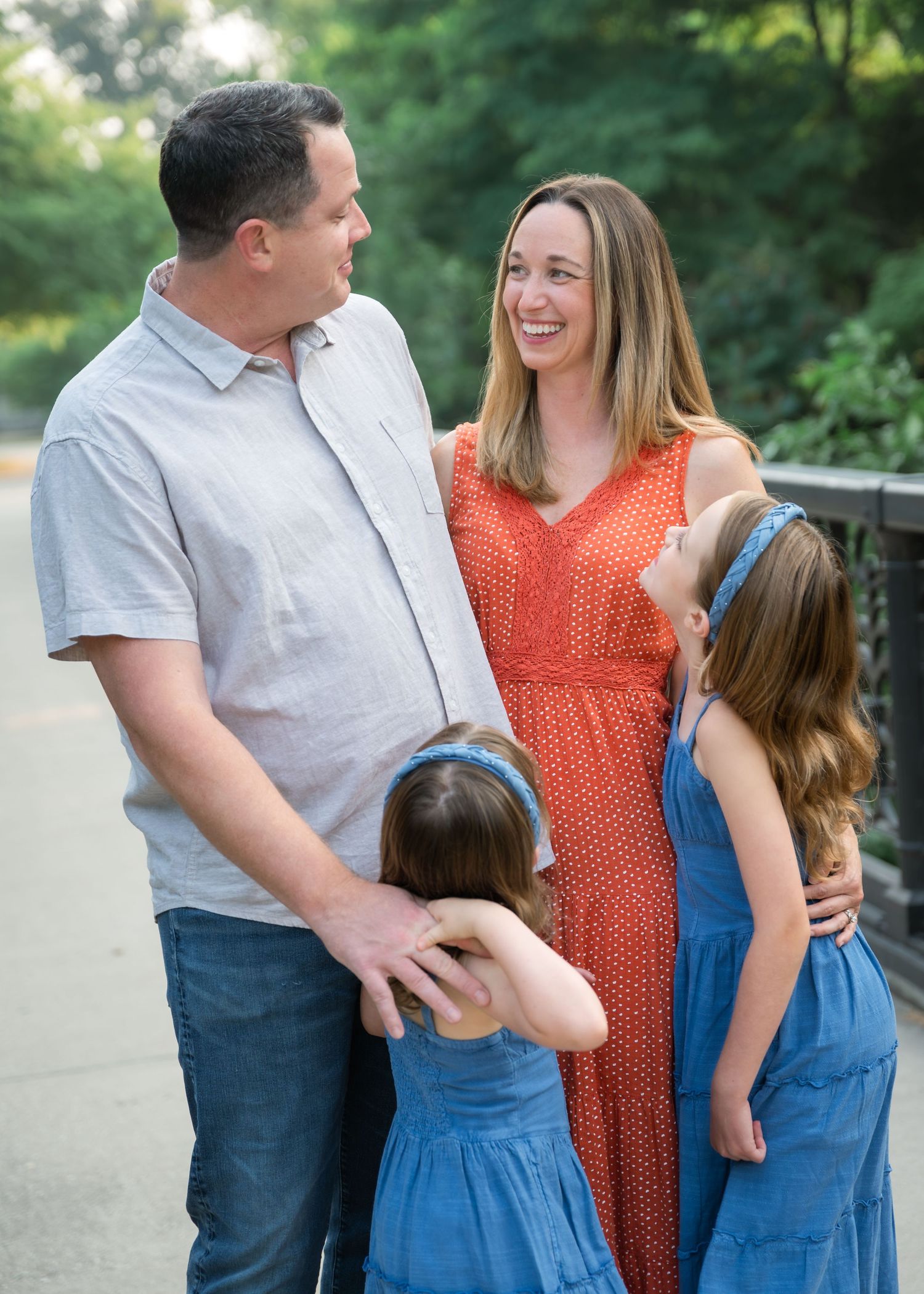 A happy family of four poses outdoors, with parents and two girls in blue dresses sharing a joyful moment.