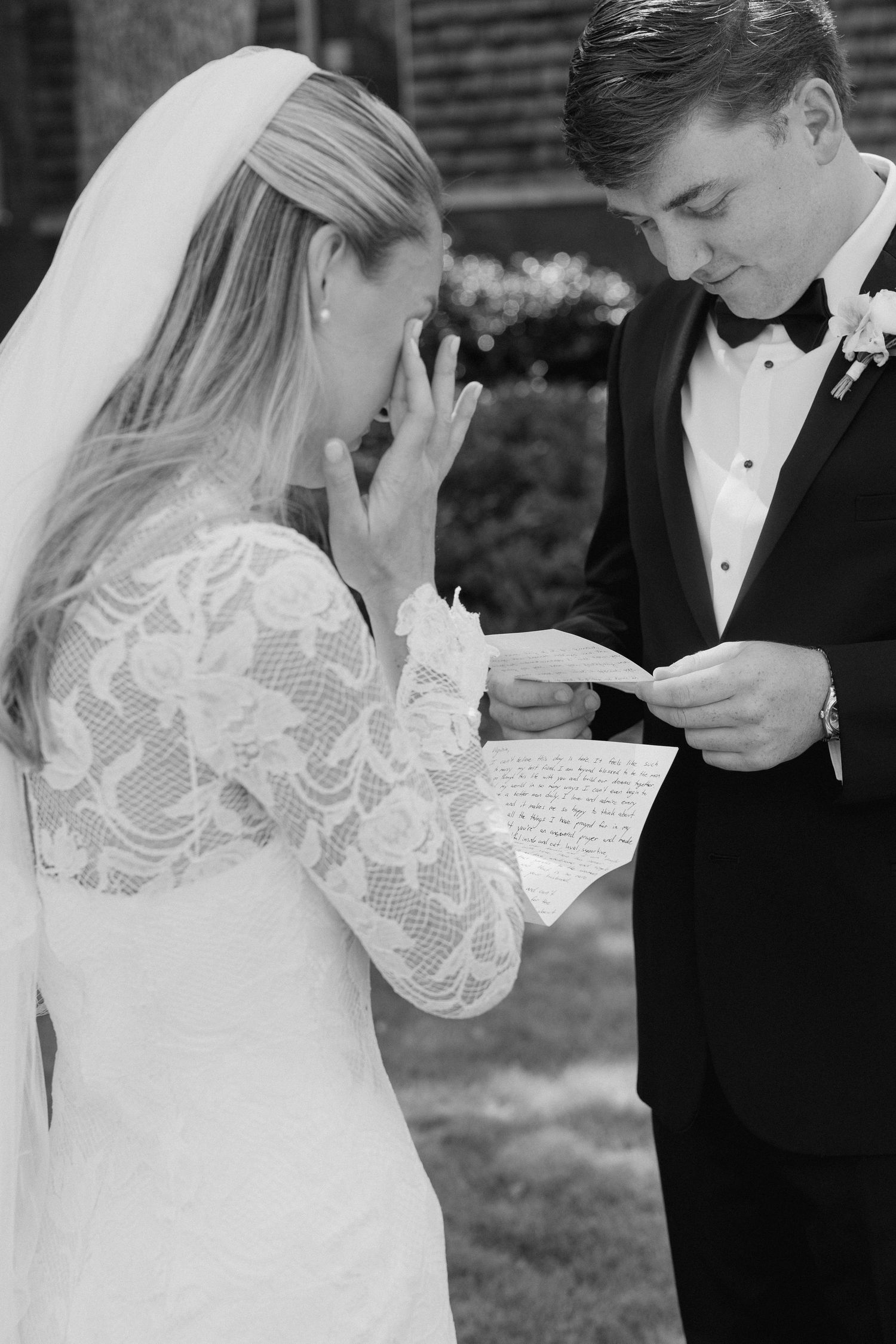 Emotional bride in lace gown wipes tears as groom reads heartfelt wedding vows from letter outdoors.