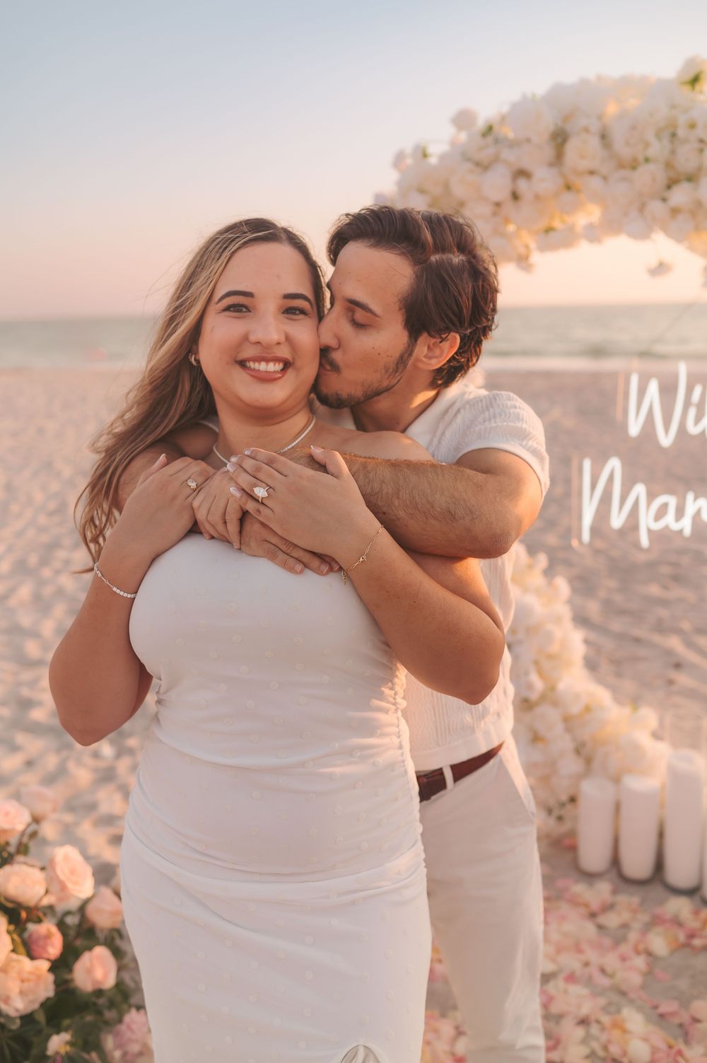 An engaged couple embraces on the beach near a floral heart arch after a romantic proposal.