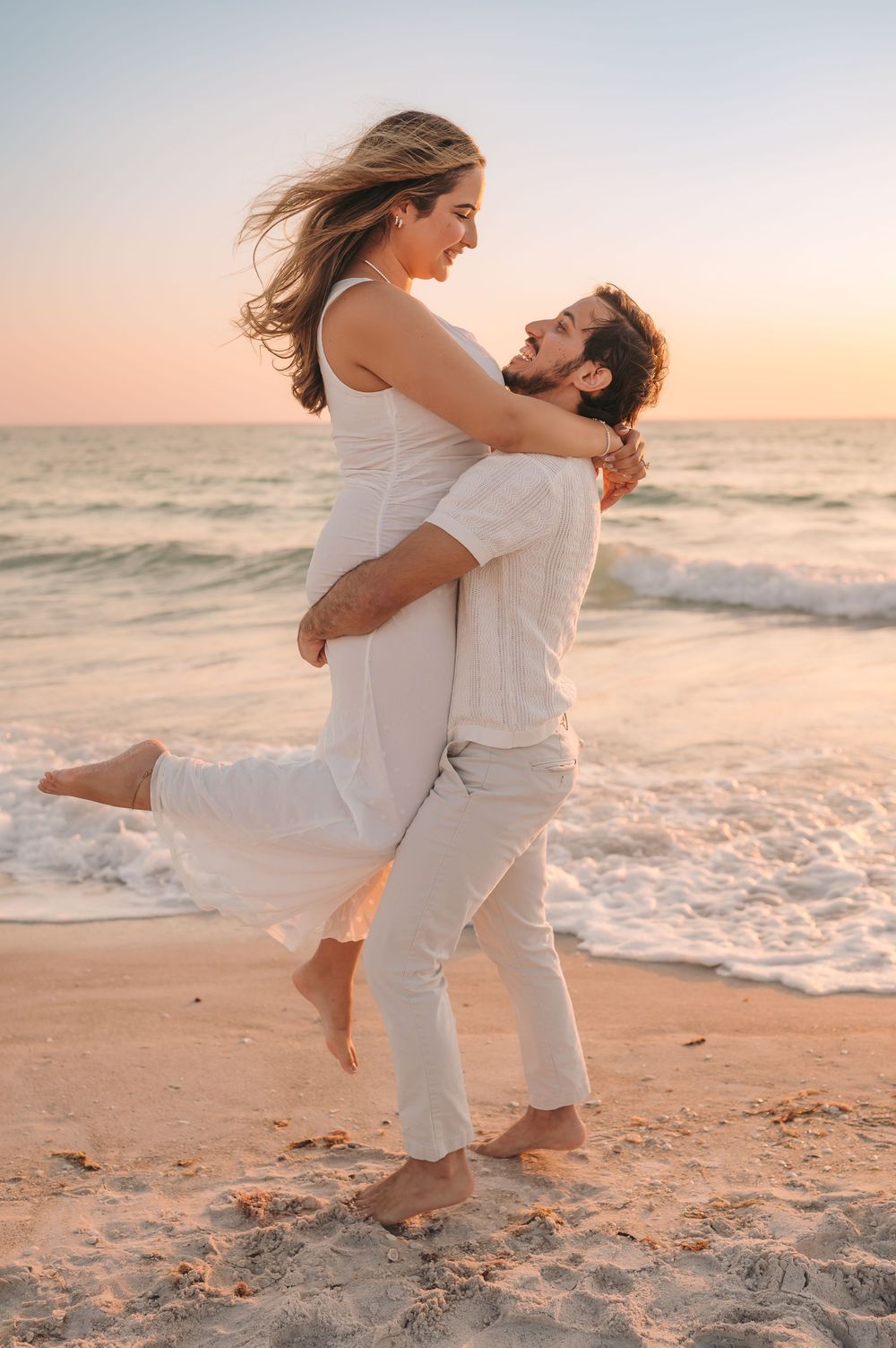 Man lifting woman in white dress on sandy beach at colorful sunset with ocean waves.