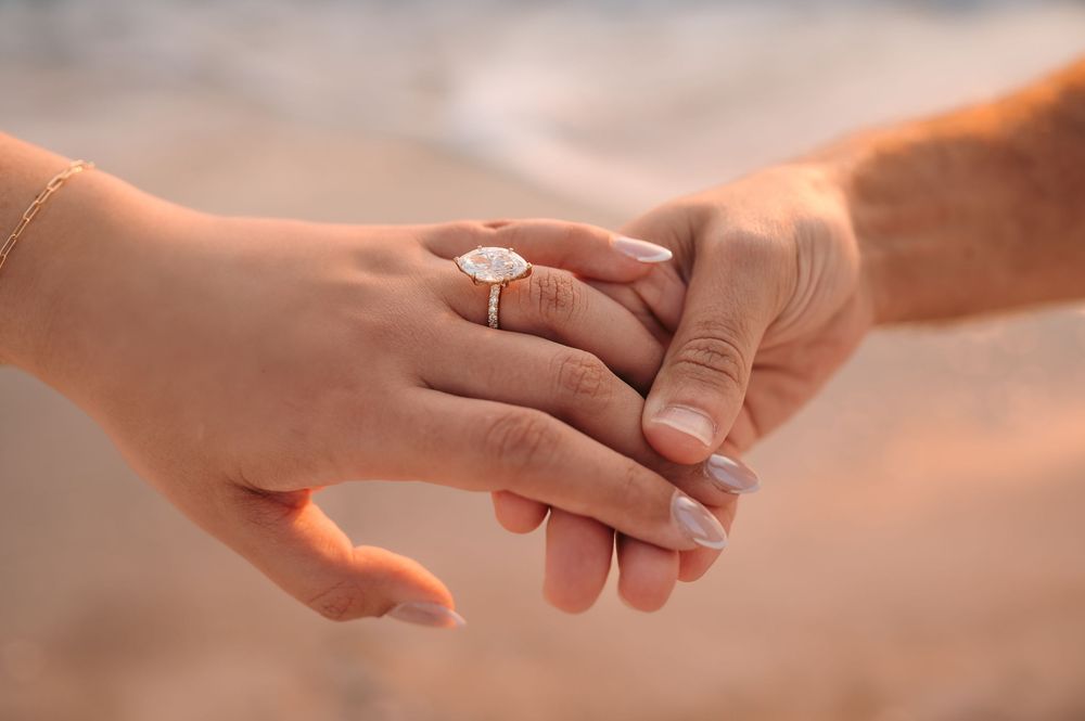 Close-up of engaged couple holding hands showing oval diamond engagement ring on beach.