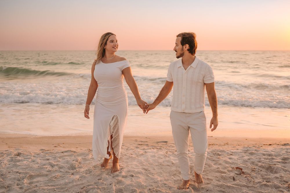 Happy couple in white outfits holding hands walking on beach at golden sunset.