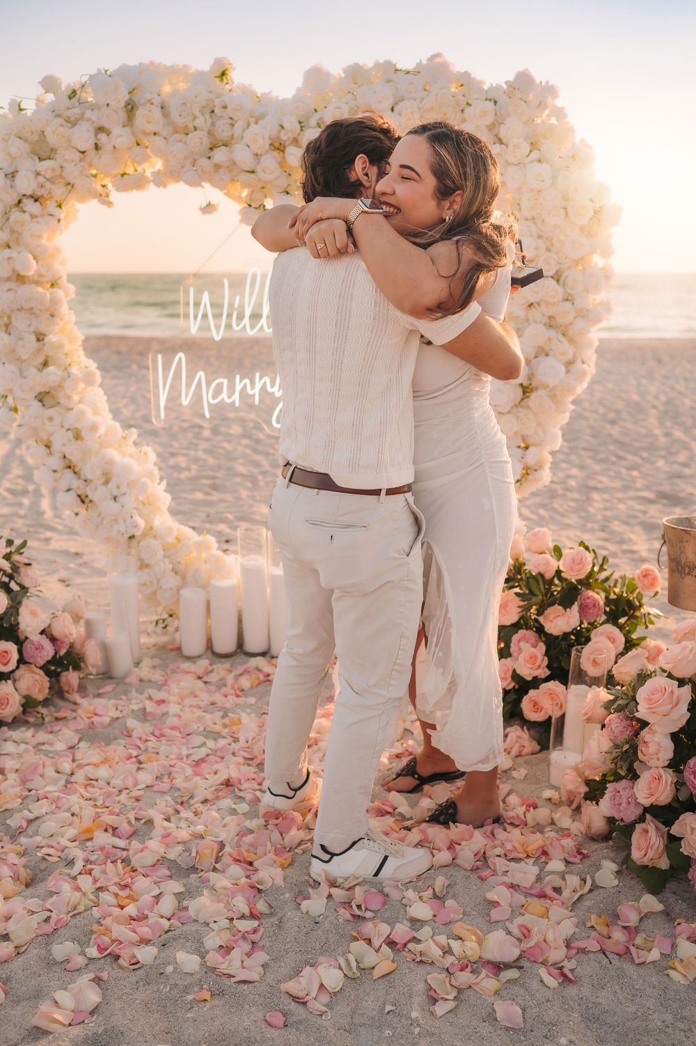 Couple hugging under heart-shaped white floral arch with Will You Marry Me neon sign on beach.
