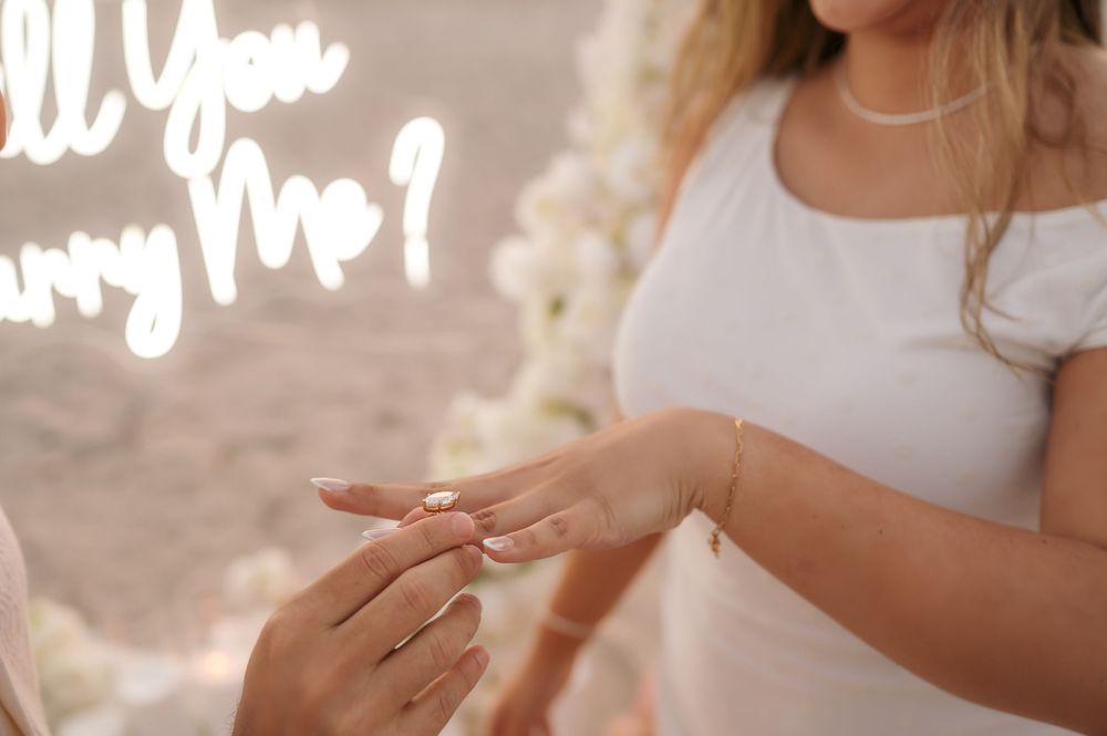 Groom placing diamond engagement ring on brides finger with Will You Marry Me neon sign backdrop.