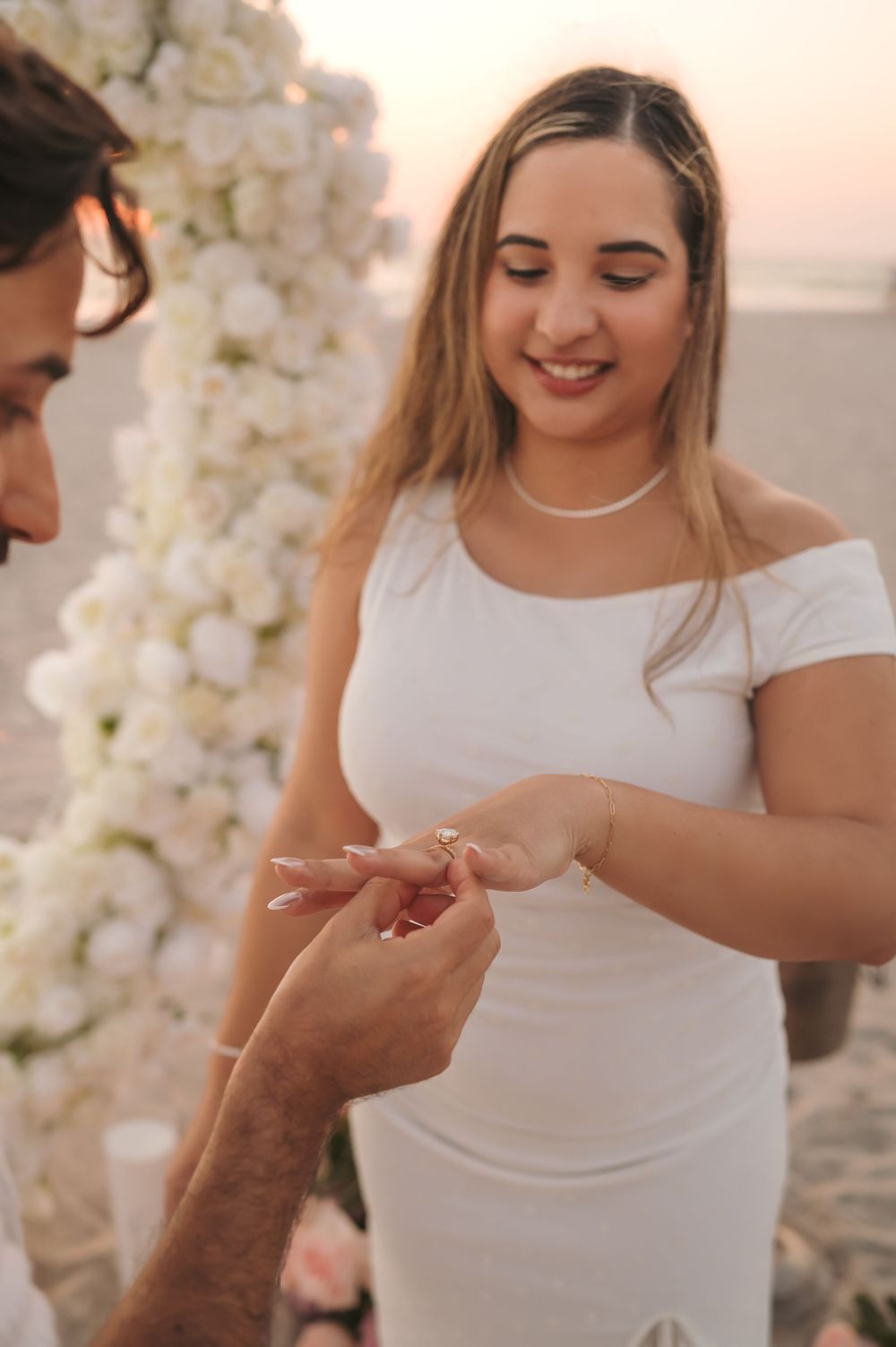 Smiling bride in white off-shoulder dress receives engagement ring from groom at beach floral arch ceremony.