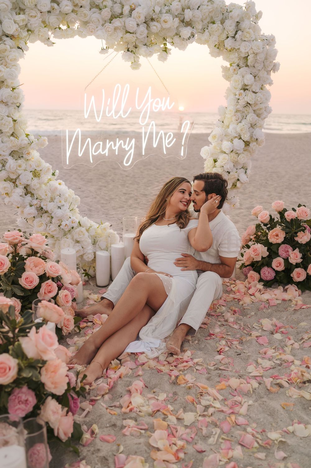 Couple embracing under floral heart arch on beach at sunset with pink roses and Will You Marry Me neon sign.