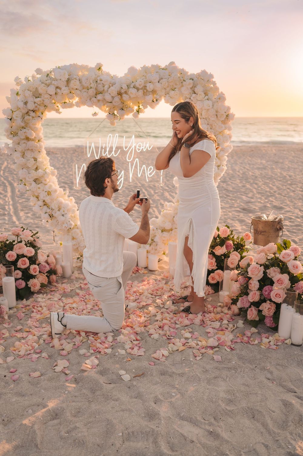 A man proposes on bended knee to his girlfriend inside a floral heart arch on a sunset beach.