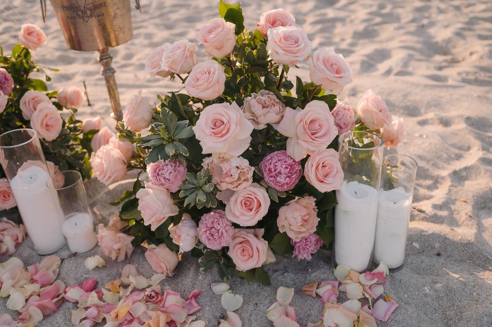 Pink roses and peonies floral arrangement with white candles and rose petals on sandy beach.