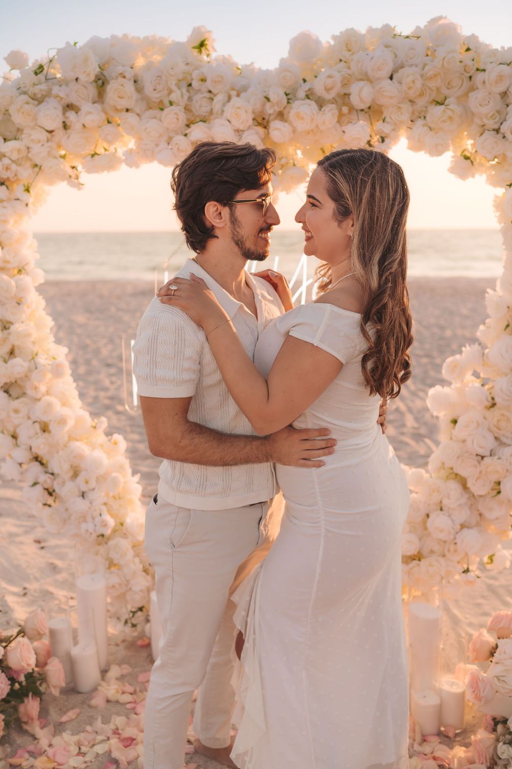 Couple embracing under white floral heart arch on beach at sunset during romantic proposal.