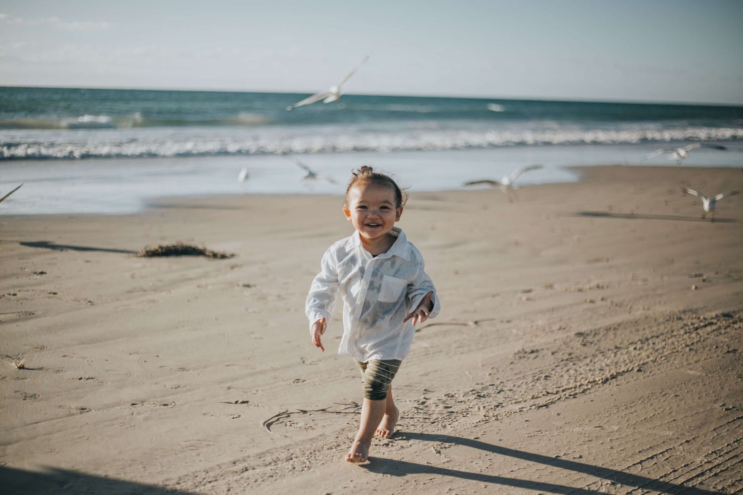 A happy toddler runs barefoot on a sunny beach with seagulls flying overhead and ocean waves in the background.