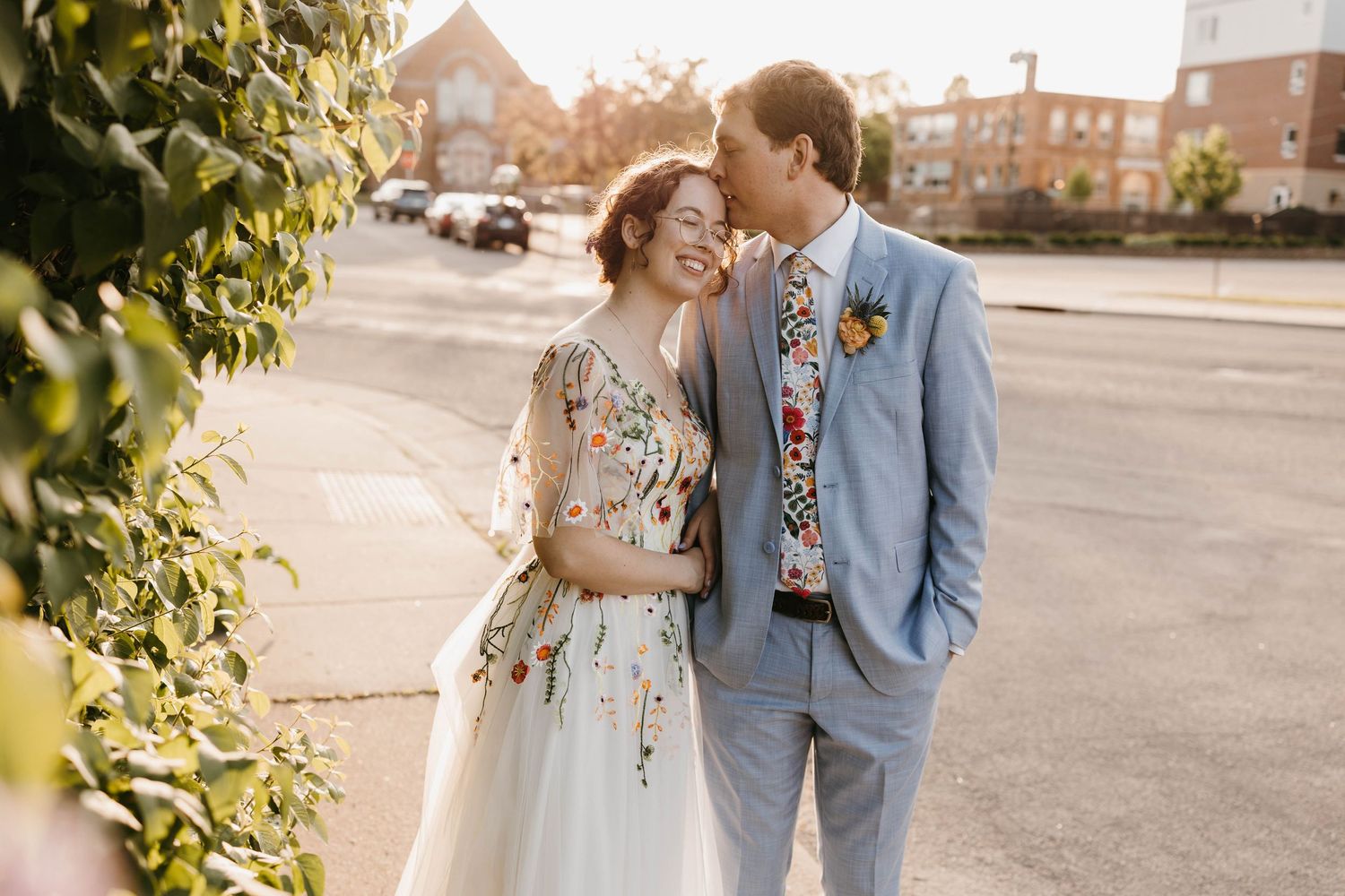 Bride in floral gown and groom in light blue suit share a tender moment outdoors on their wedding day.