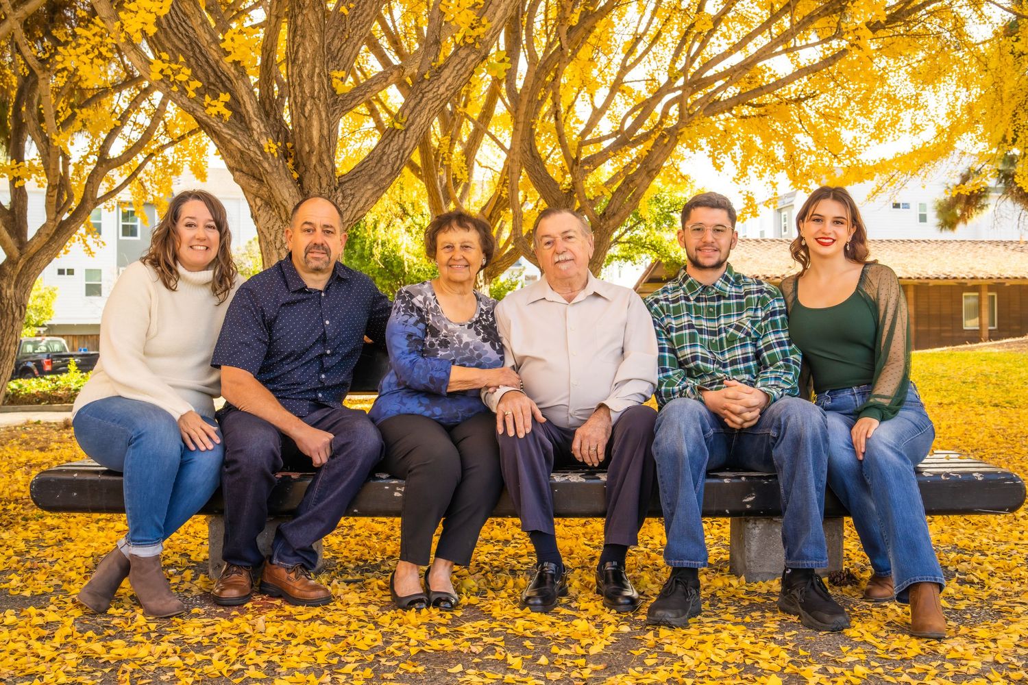 Six family members sitting on a bench under golden autumn trees with fallen yellow leaves on the ground.
