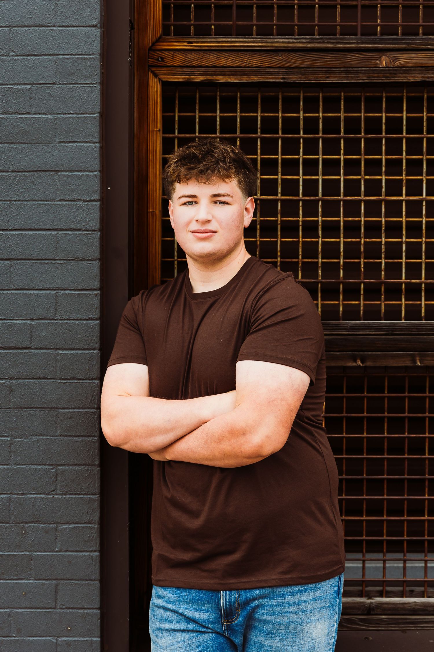 Young man in brown t-shirt and jeans stands confidently with arms crossed against urban brick wall backdrop.