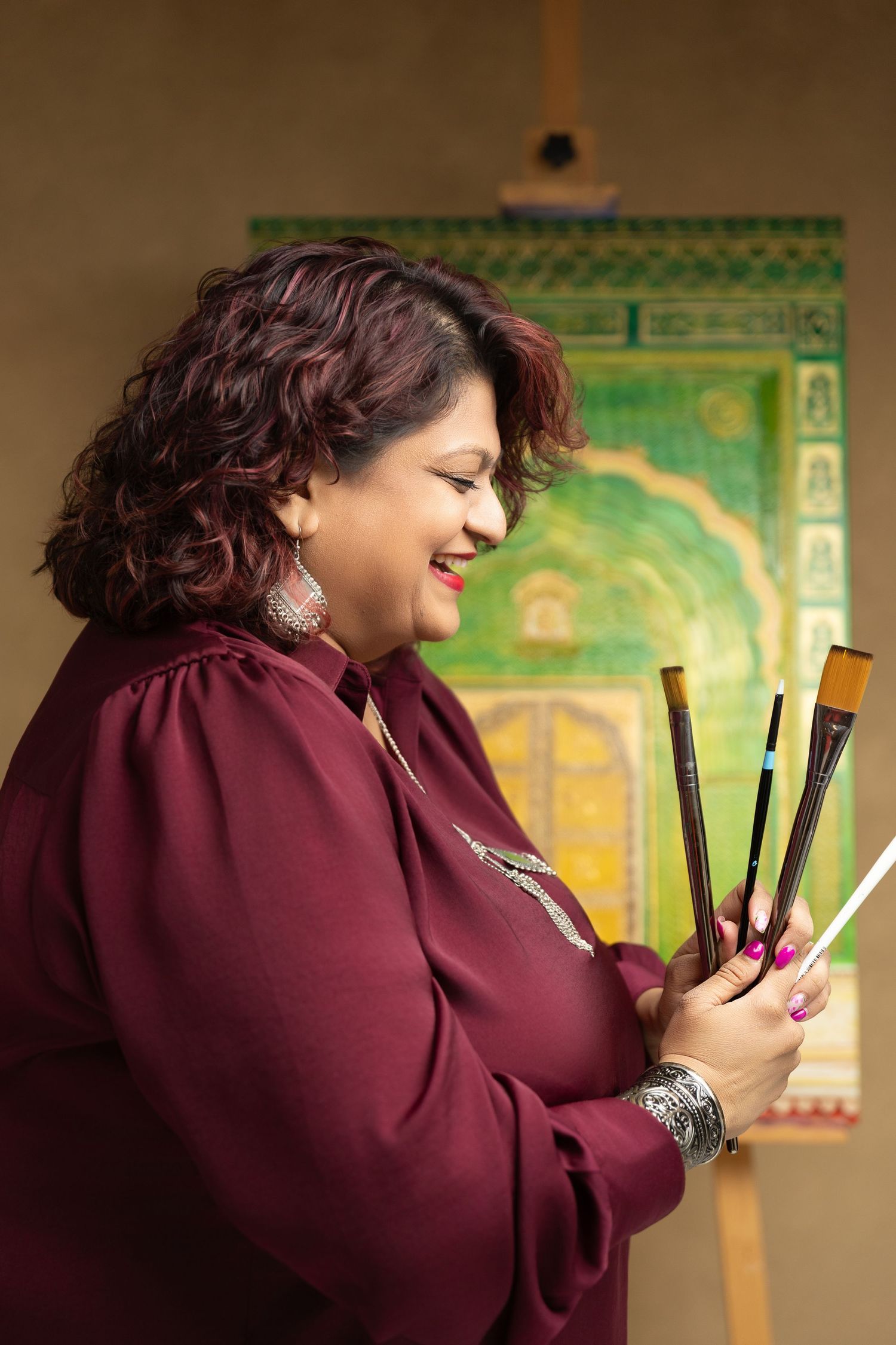 A smiling woman in burgundy holds paint brushes, standing before a colorful green architectural painting on canvas.