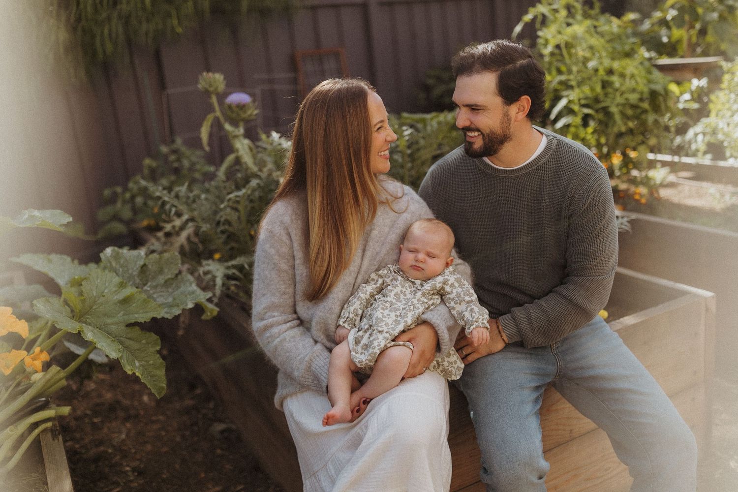 A happy couple sitting outdoors in a garden, smiling at their newborn baby held in the mother's arms.