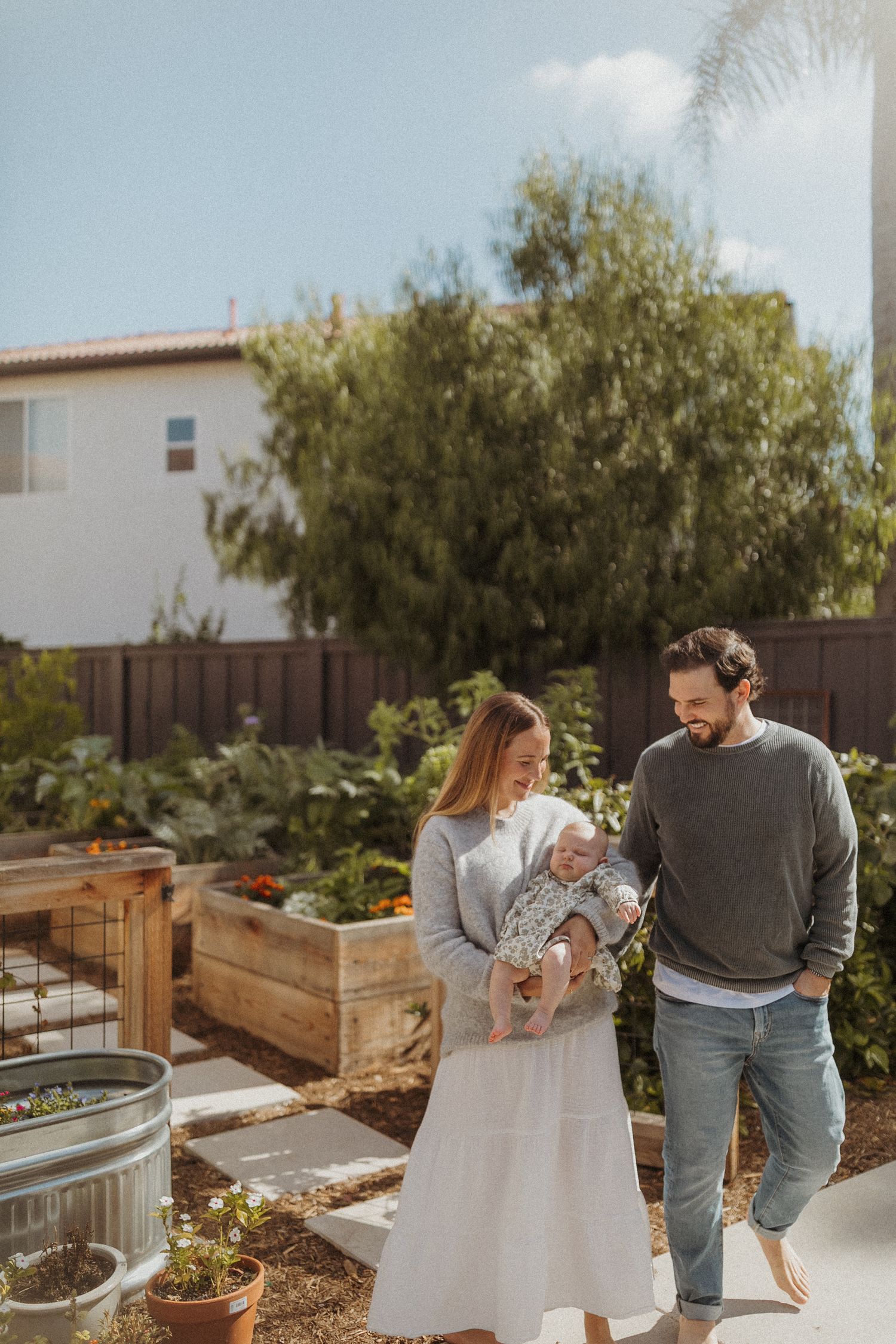 A couple walks through a backyard garden with raised beds, the woman holding a baby while the man smiles at them.