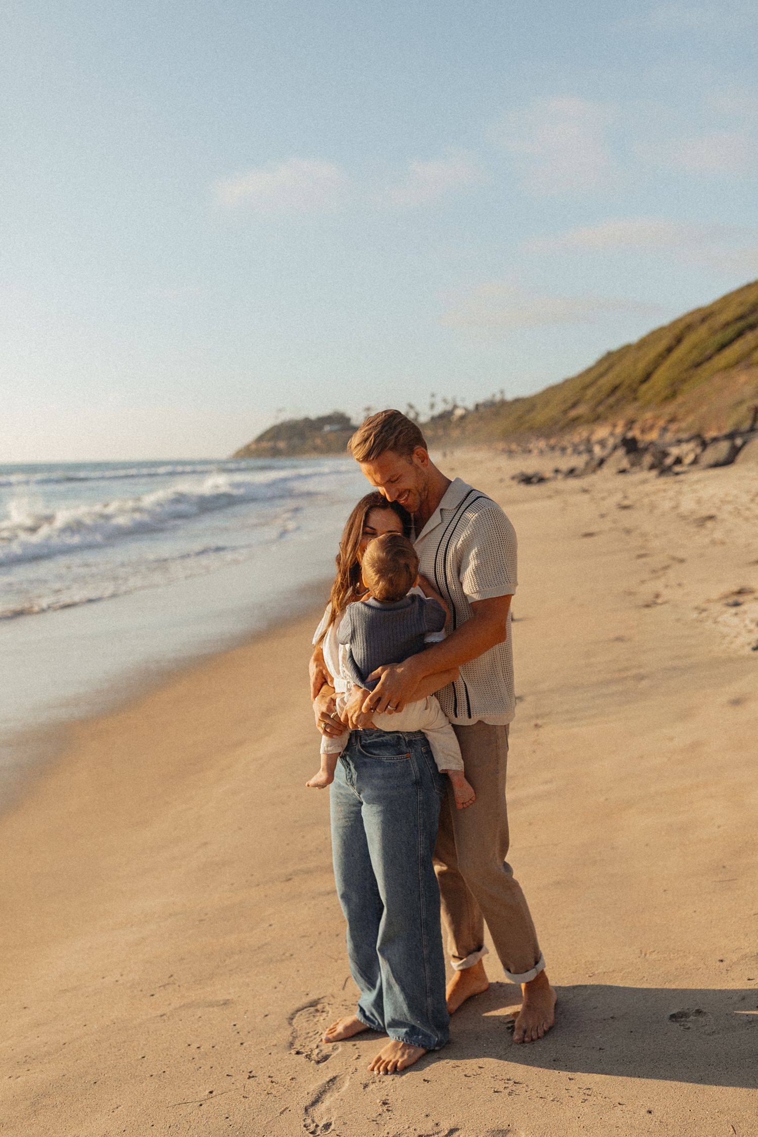 A couple embraces tenderly on a sunny beach, holding a baby, with ocean waves and a green hillside in the background.