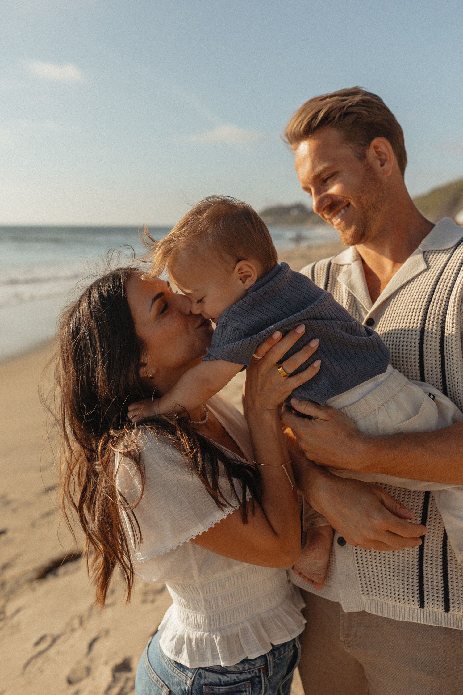 A happy young family with a baby enjoys a sunny beach day, with the mother kissing her infant while the father smiles warmly.