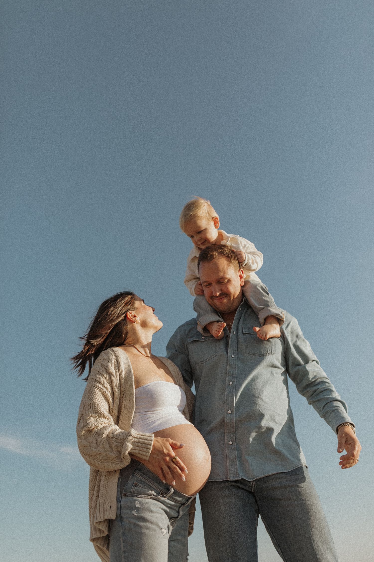 A happy pregnant woman and man with toddler on shoulders pose outdoors against a clear blue sky.