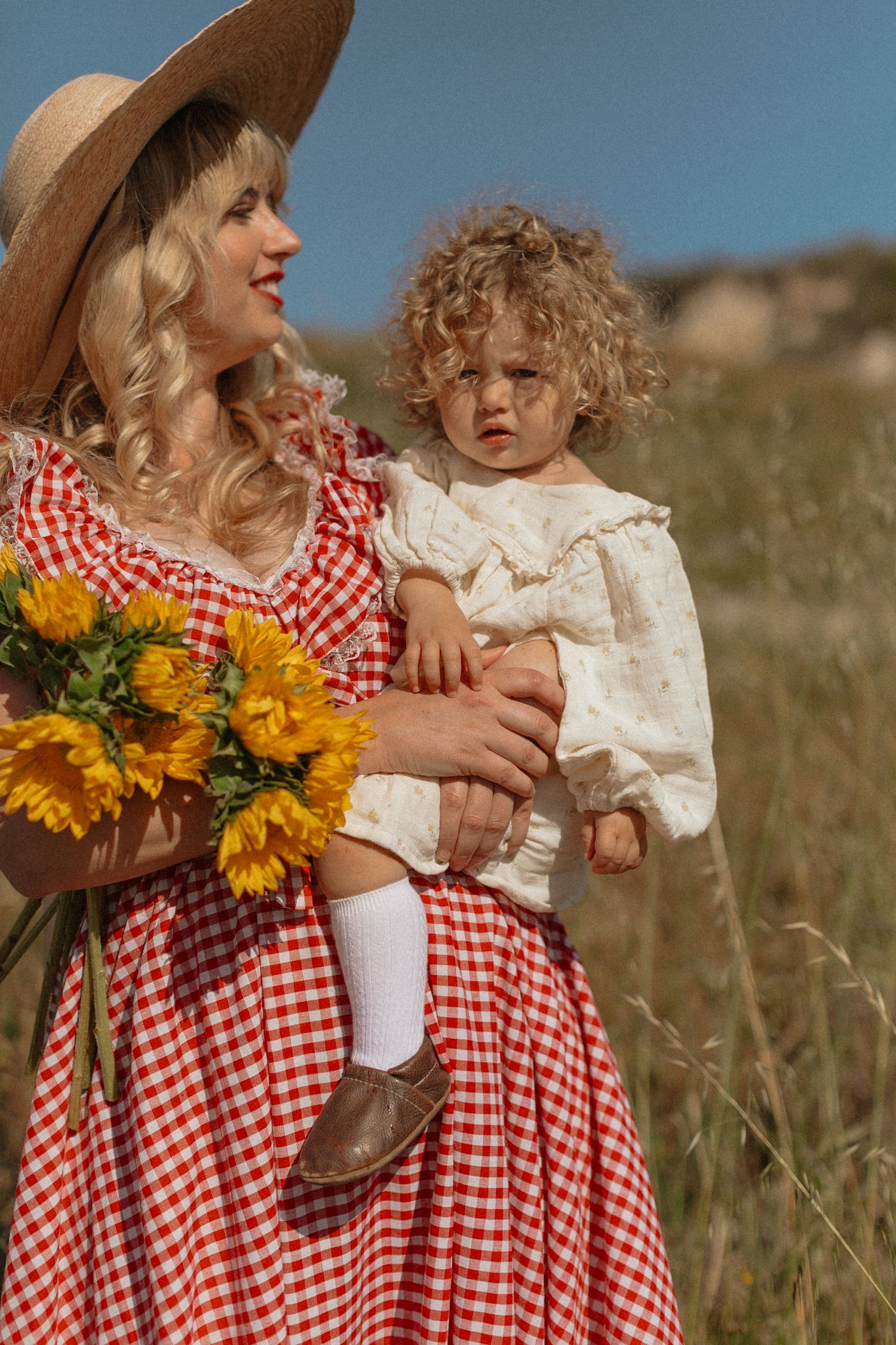 A woman in a red gingham dress and straw hat holds a curly-haired toddler and sunflowers in a sunny field.