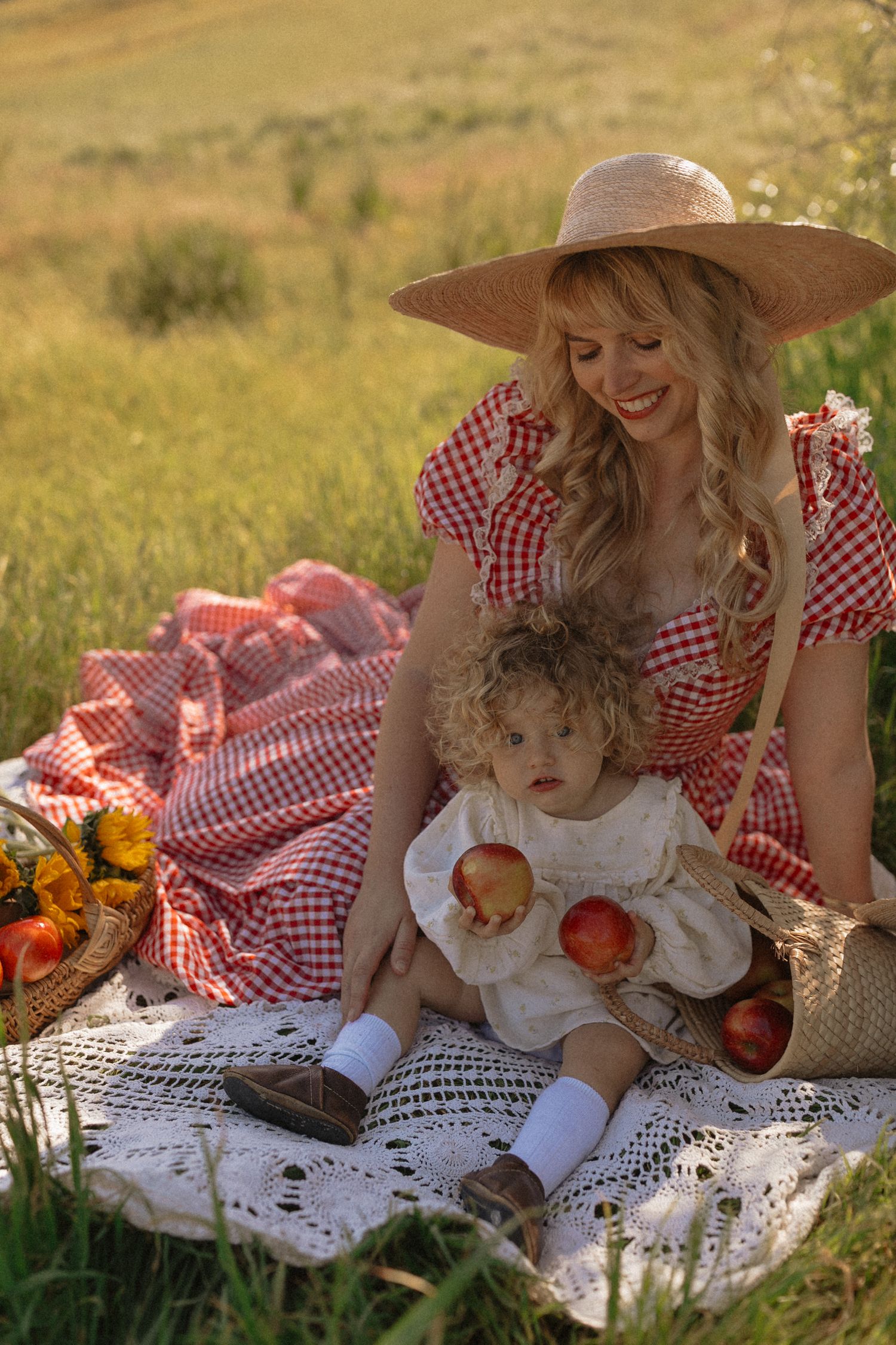 Mother in red gingham dress and straw hat sits with toddler on lace blanket during sunny outdoor picnic with apples.