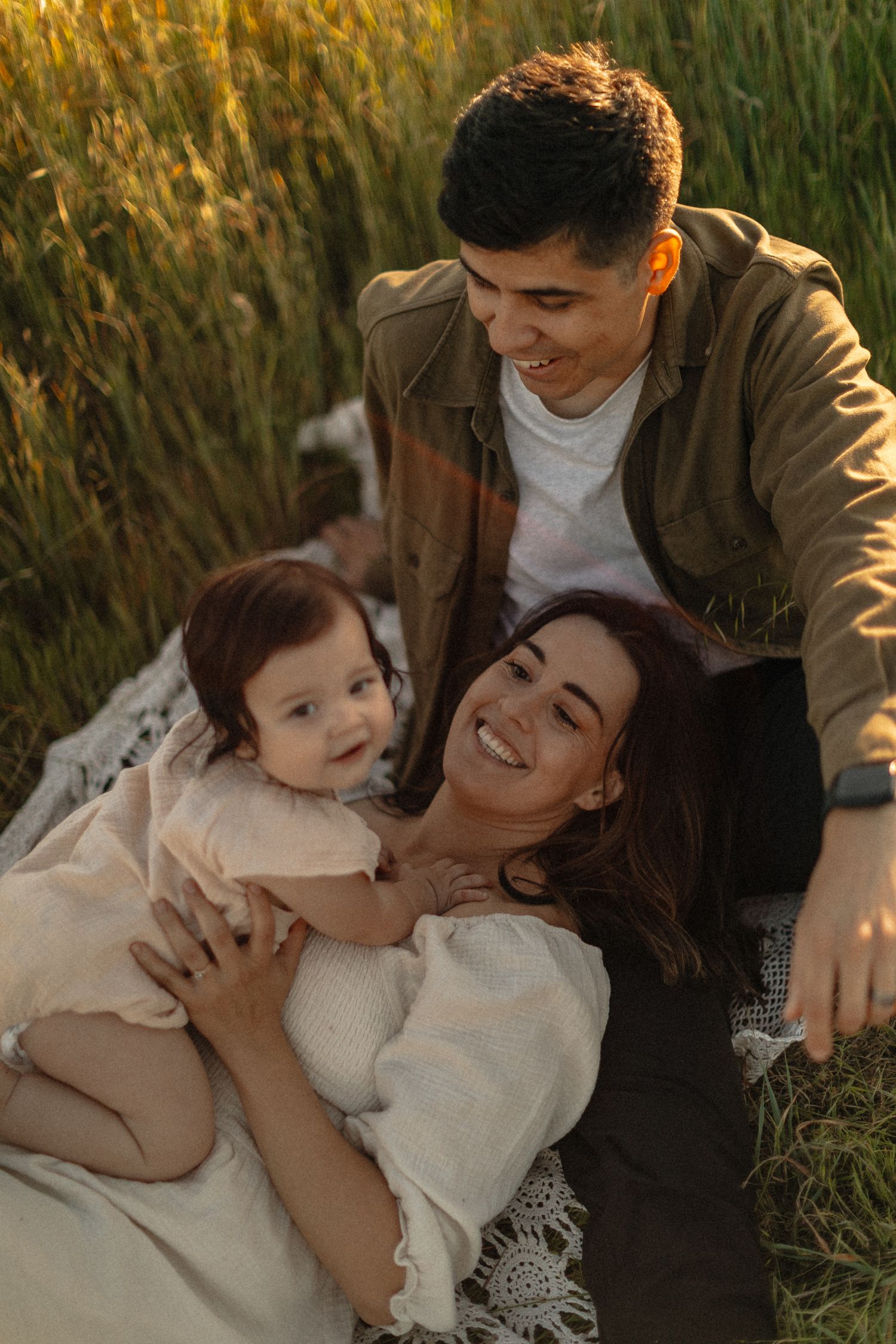 Happy young family with baby relaxing on blanket in green field during golden hour sunset.