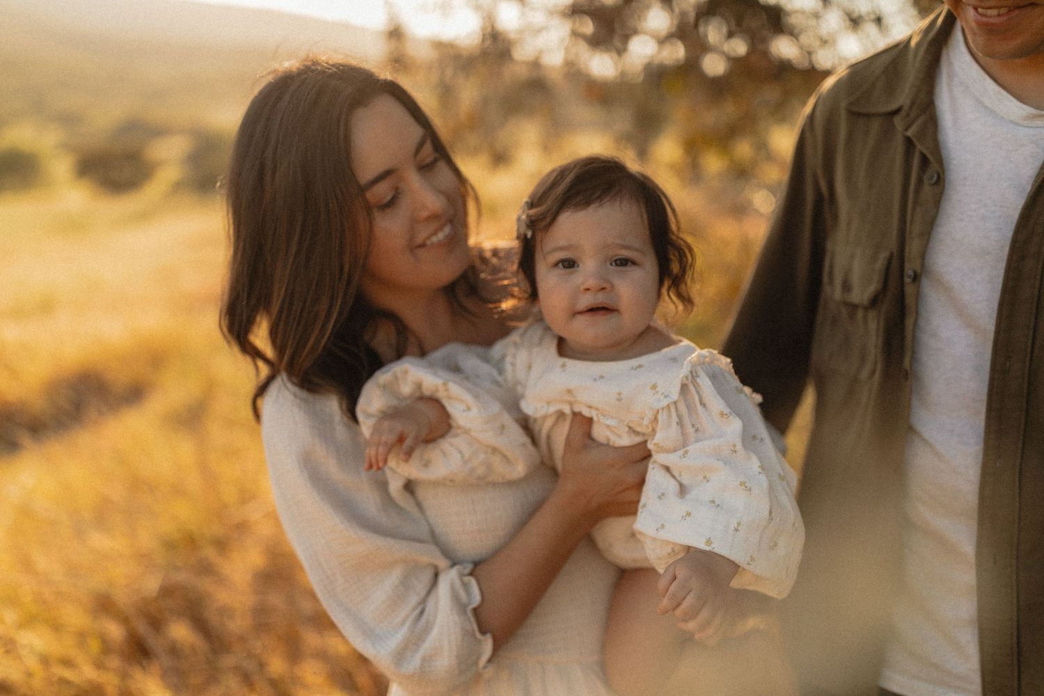 A mother holds her baby girl in a golden field at sunset, with a man standing beside them.