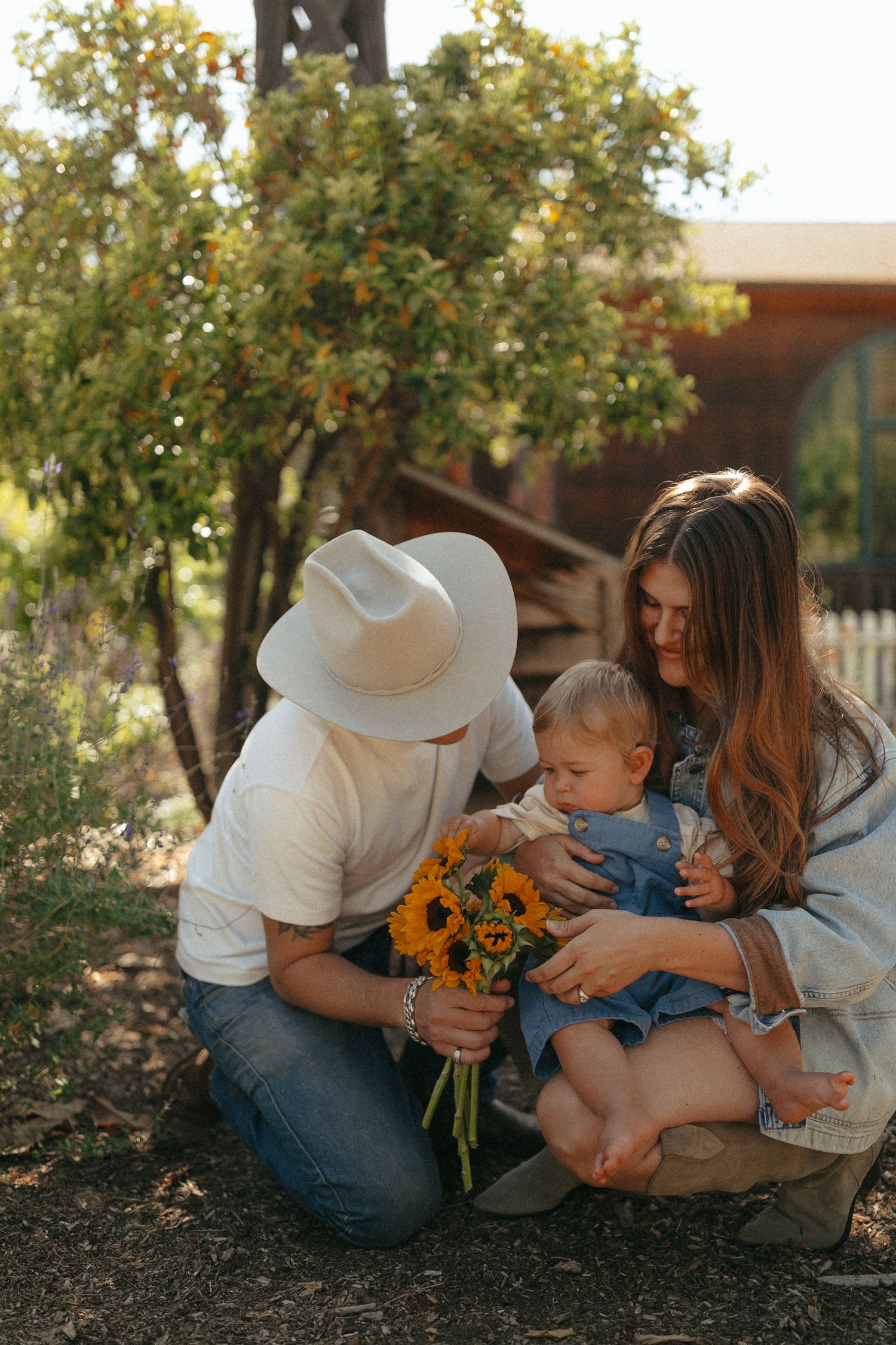 A couple crouches outdoors with their baby, sharing a bouquet of sunflowers near a lush green tree.