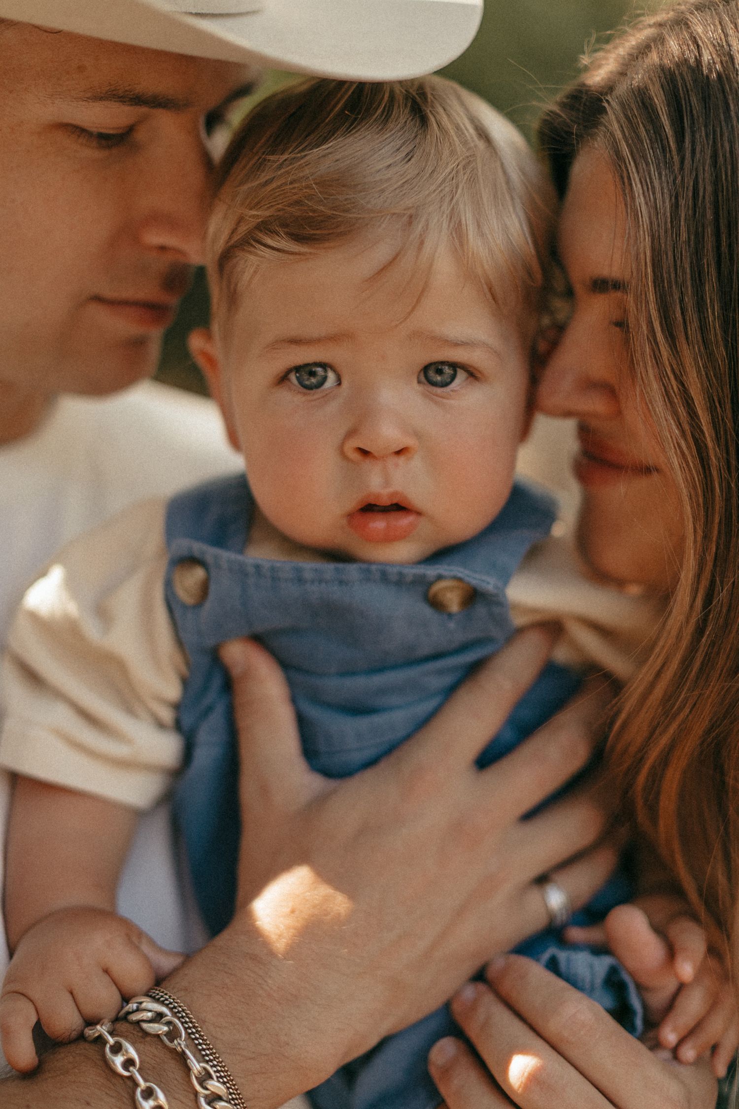 Parents lovingly hold their blue-eyed baby dressed in denim overalls in a warm, sunlit outdoor setting.
