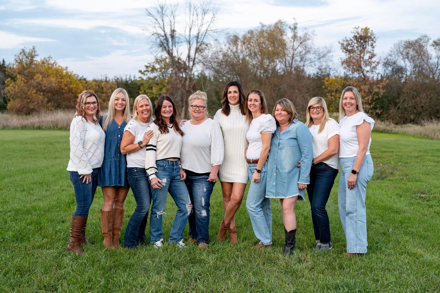 Group of nine women smiling outdoors in a green field, wearing casual white and blue outfits on a fall day.