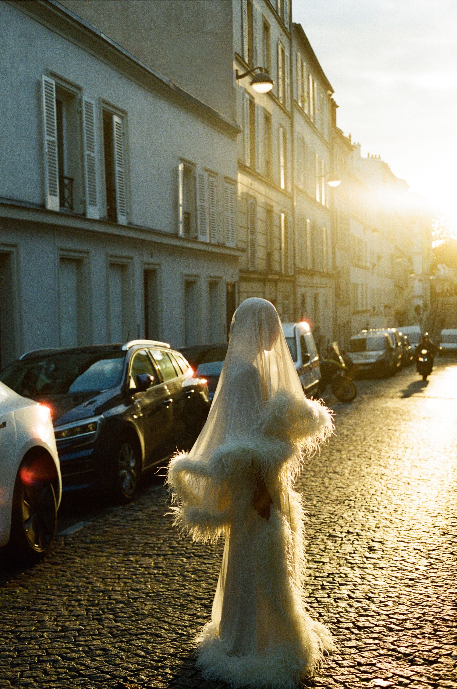 A figure in a white feathered gown and long veil walks a golden sunlit cobblestone Paris street at sunset.