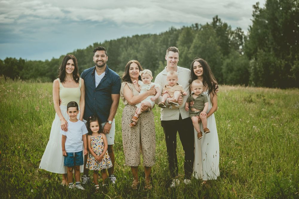 Large family group portrait in a green meadow with tall trees, featuring adults and young children outdoors St Albert Family Photographer