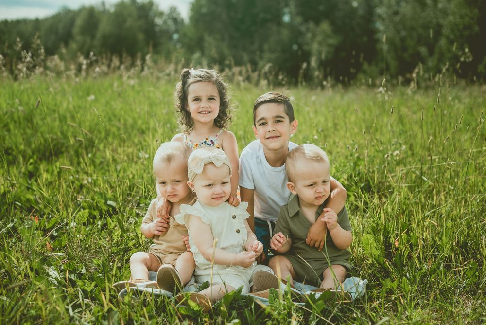 Five young children smiling and sitting together in a lush green meadow on a sunny day. St Albert Family Photographer
