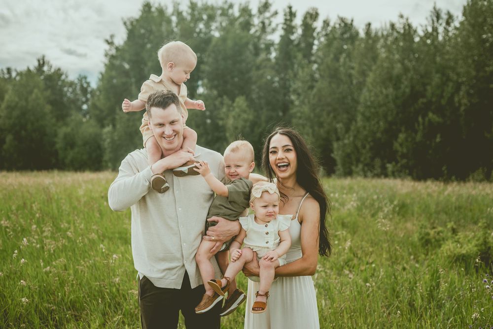 Happy family with three young children posing in a lush green meadow with trees in the background. St Albert Family Photographer