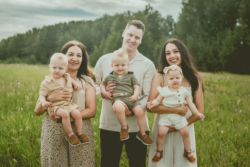 Family of five posing outdoors in a green field, parents holding three young babies, smiling on a cloudy day. St Albert Family Photographer