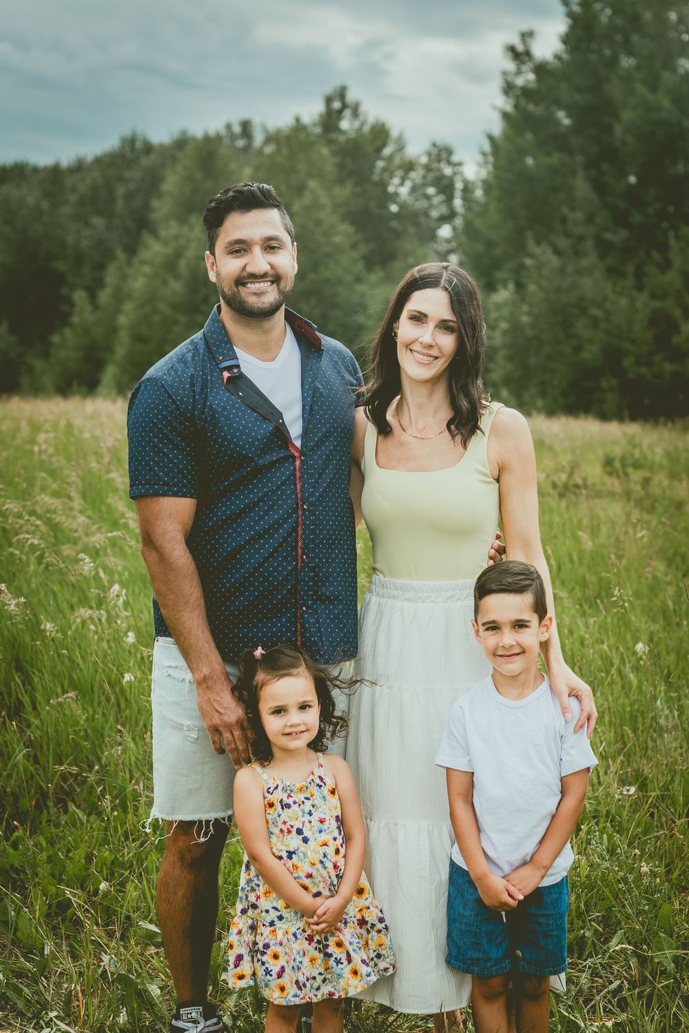 Family of four smiling outdoors in a green meadow, parents with two young children on a cloudy day. St Albert Family Photographer