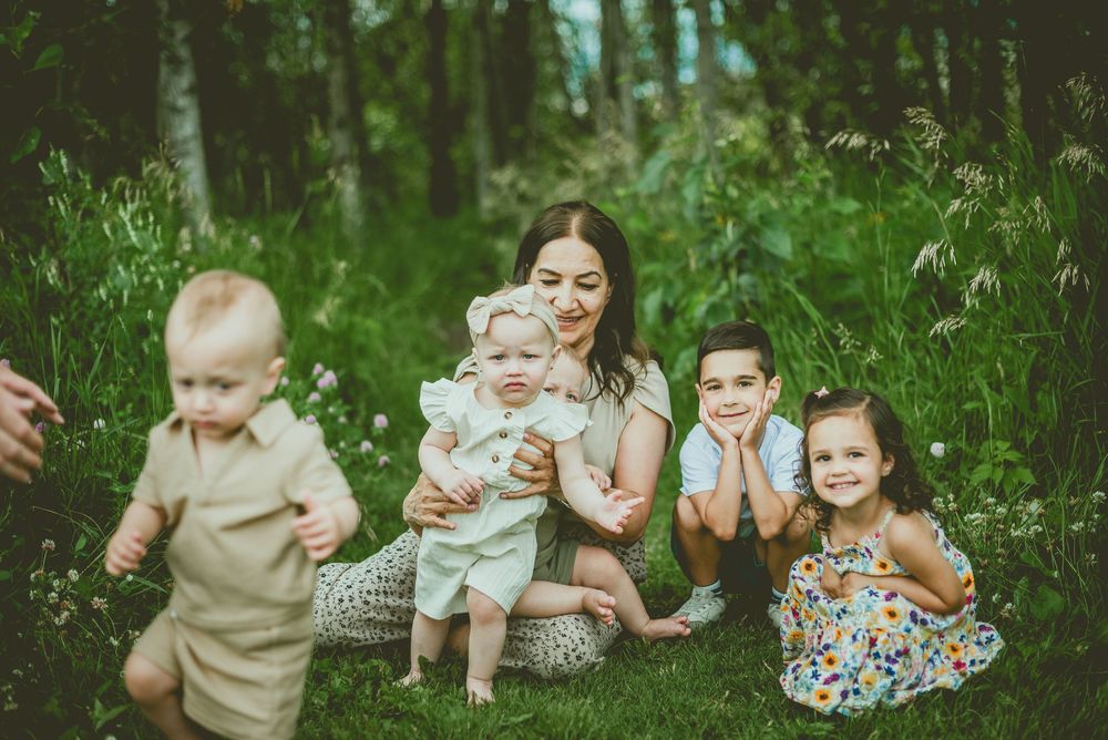 A smiling mother sits on lush green grass with young children, including a baby, in a serene forest setting. St Albert Family Photographer