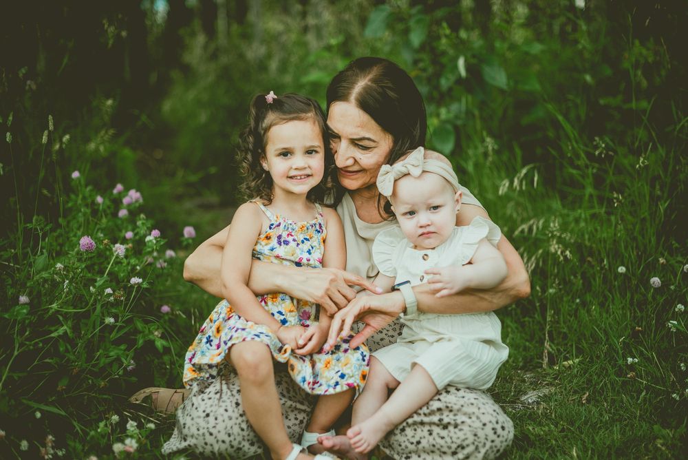 A mother sits outdoors in lush greenery, embracing her two young daughters in a heartwarming family portrait. St Albert Family Photographer