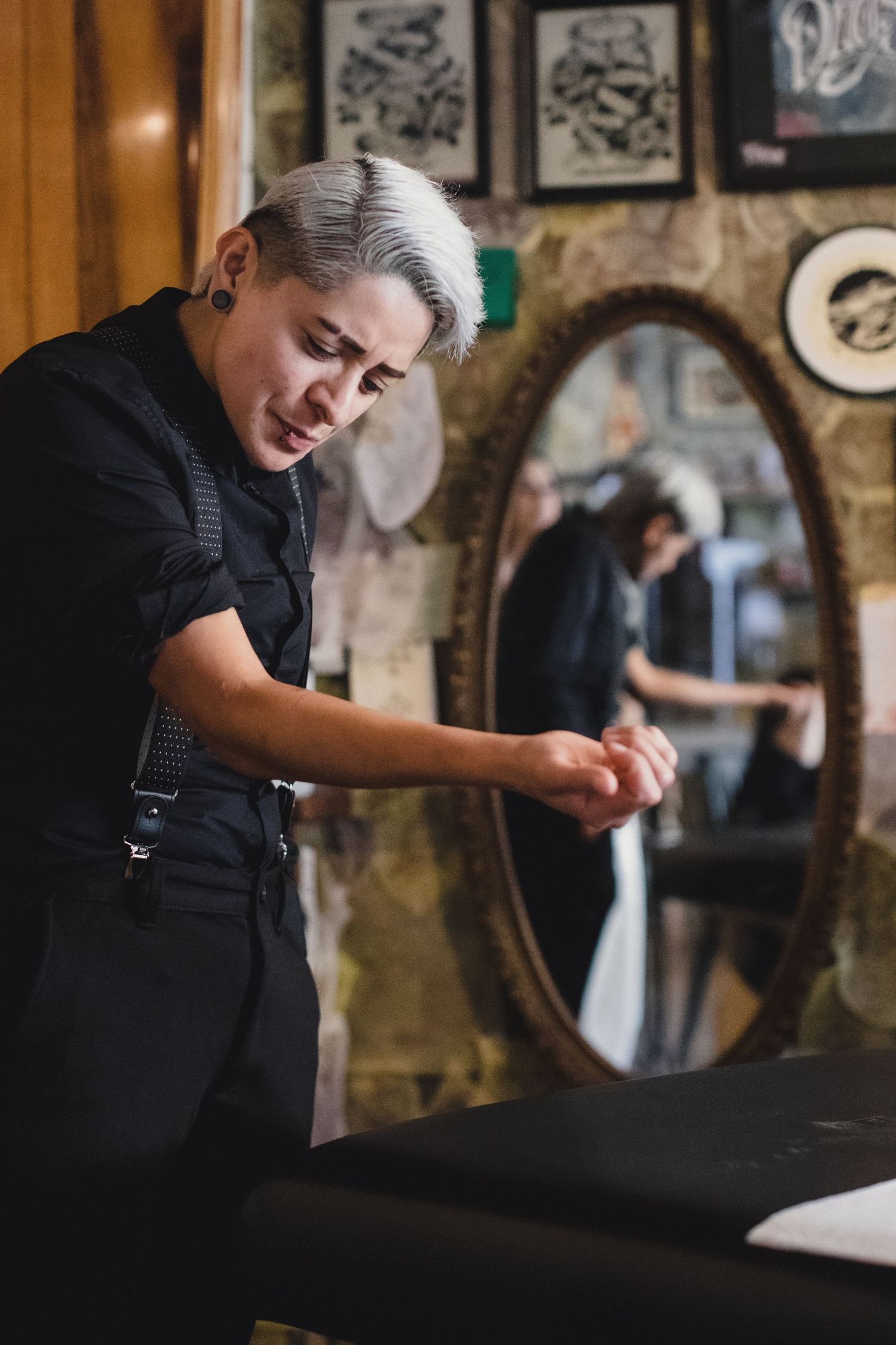 Short-haired tattoo artist in black outfit preparing hands in a decorated tattoo studio with mirror reflection.
