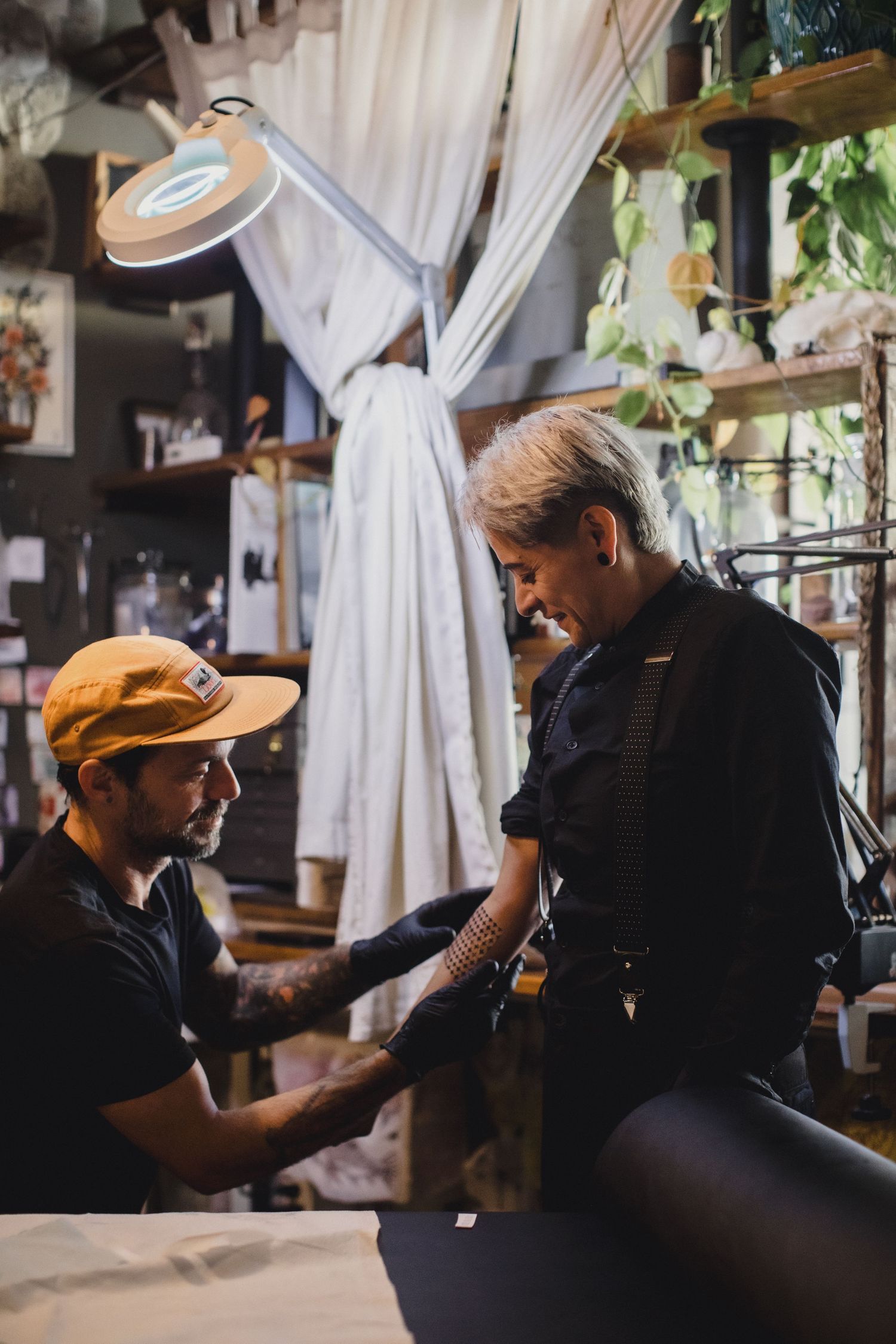 Tattoo artist in orange cap applying tattoo on client's arm in a decorated studio with warm lighting.