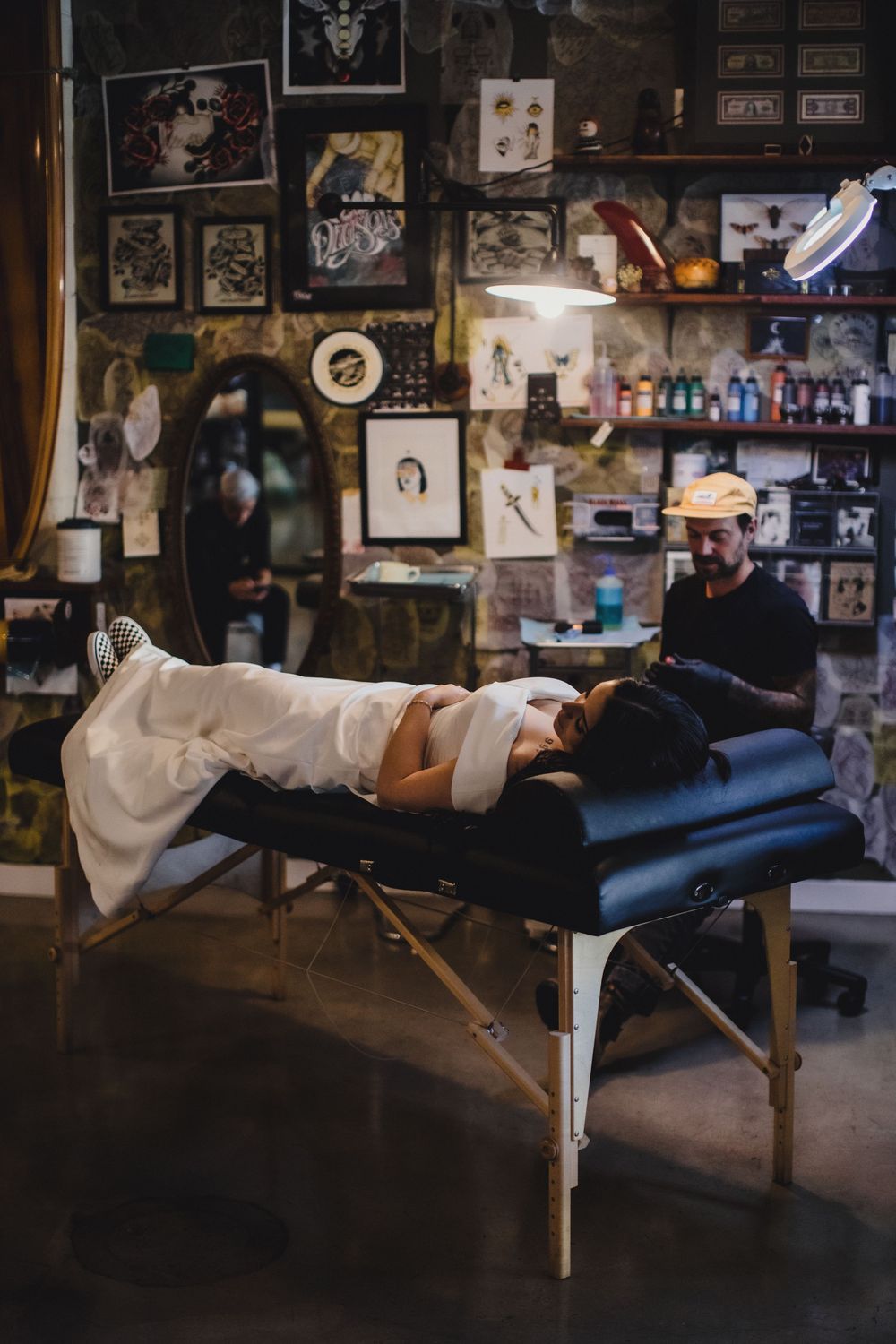 A tattoo artist works on a client lying on a black massage table in a decorated studio with artwork on walls.