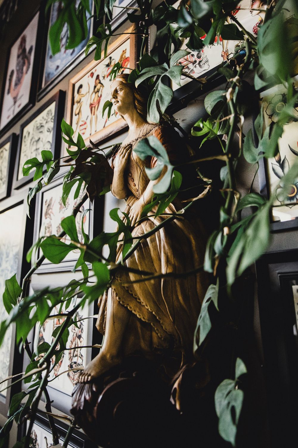 A classical female statue surrounded by lush green vines, with framed artwork displayed on a dark wall behind.