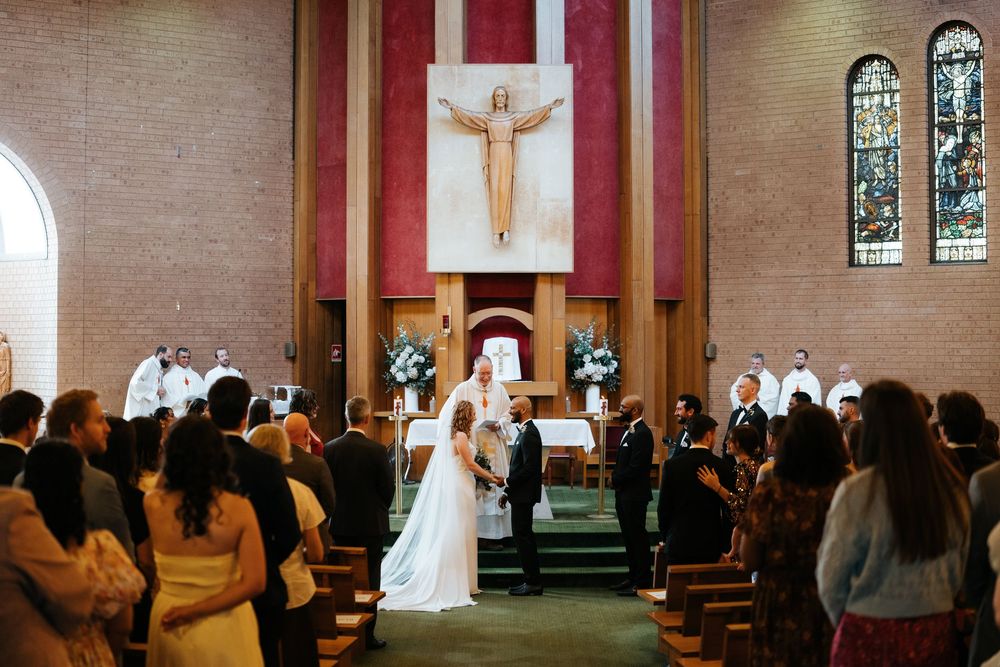 A bride and groom exchange vows at a Catholic church wedding ceremony with guests seated in wooden pews.