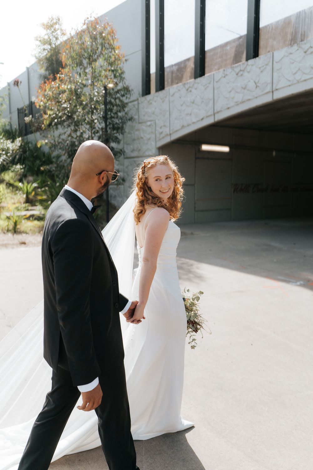 Bride in white gown holds groom's hand while walking outdoors, glancing back with a smile on their wedding day.