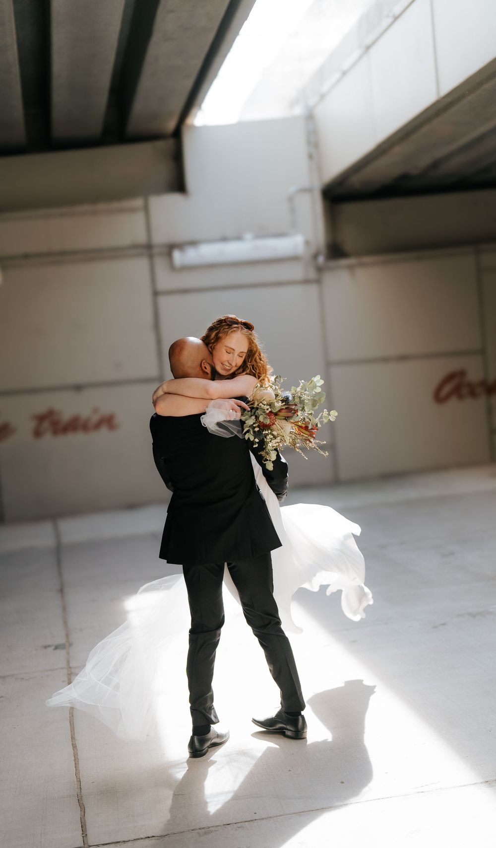 Bride and groom embracing in a sunlit urban venue, bride holding a wildflower bouquet with veil flowing.
