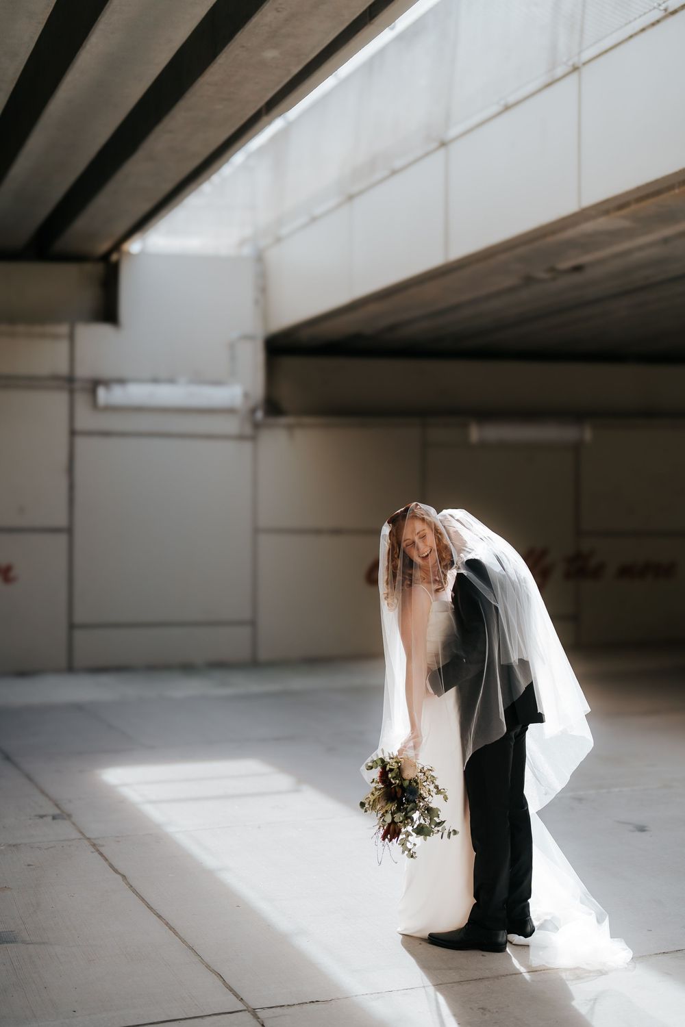 Bride and groom share a romantic kiss under an urban overpass, bride holding a wildflower bouquet in natural light.