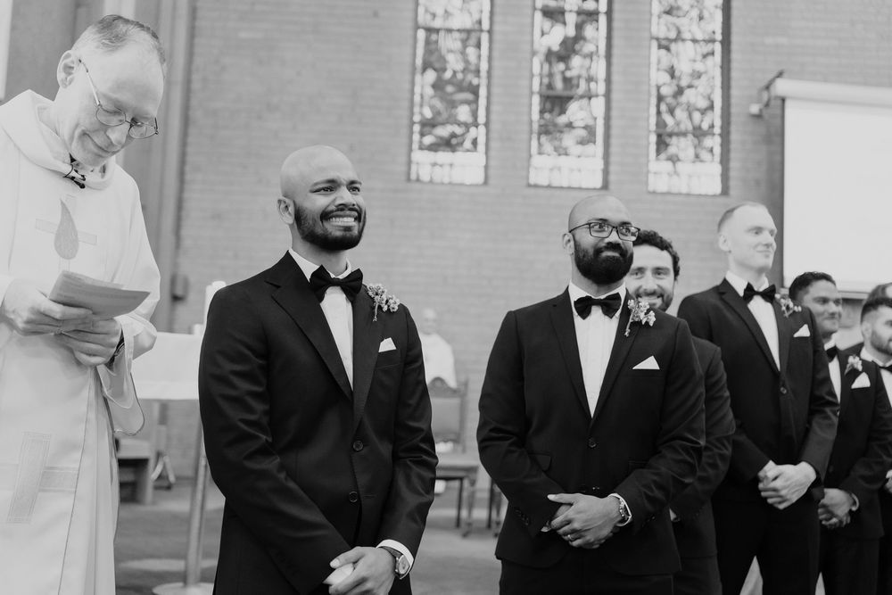 Groom and groomsmen smiling during church wedding ceremony in classic black tuxedos with bow ties and boutonnieres.